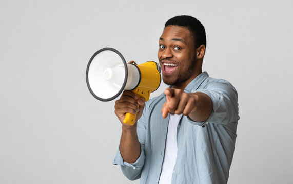 Positive african american guy shouting in megaphone and pointing at camera over grey background, copy space. Happy black man screaming with loudspeaker, cheering up, making advertisement