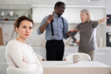 Fototapeta premium Angry middle-aged woman sitting at table while two male and female family members disputing with each other in the back