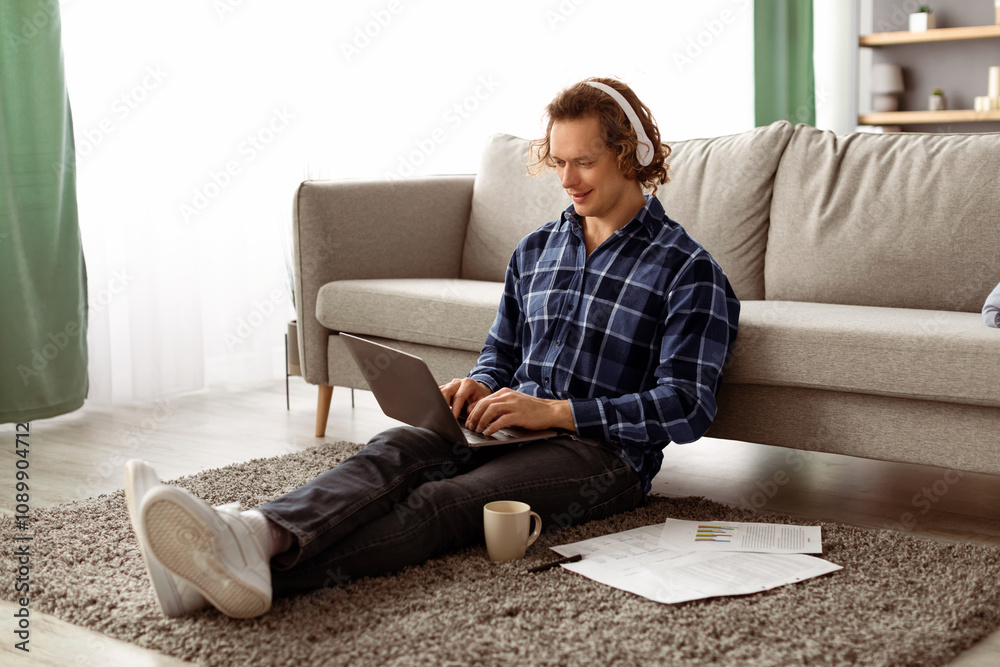 Poster happy man working on laptop wearing headphones browsing internet sitting on floor at home. freelance