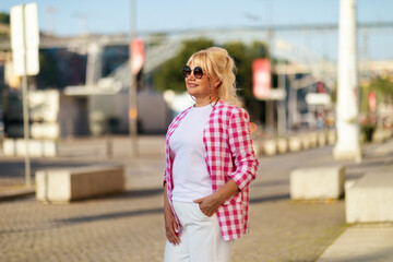 A woman stands confidently in a pink checkered shirt and sunglasses, basking in the sunlight at a bustling city plaza. The vibrant atmosphere showcases urban life and leisure.