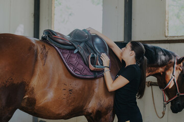 A young woman gently places a saddle on a horse inside a sunlit stable, preparing for a ride. The...