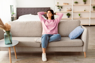 Fototapeta premium Rest Concept. Beautiful lady sitting on comfortable couch at home in living room, looking aside at window. Happy woman relaxing on sofa, leaning back, enjoying weekend free time or break from work