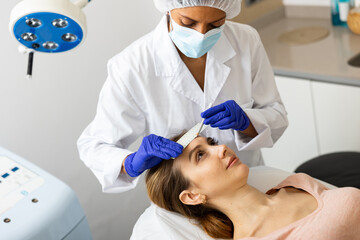 Naklejka premium Latin woman cosmetologist in face mask marking skin of young caucasian woman with white pencil and using ruler.