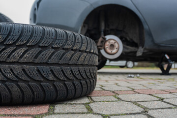 Seasonal tire replacement. A close-up of a car's removed wheel and a winter tire in the foreground. The car is lifted on a jack, showcasing the repair process taking place outdoors.