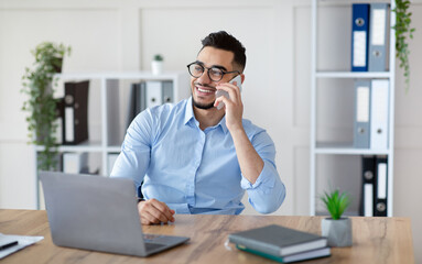 Handsome young Arab man in formal wear talking on smartphone, enjoying conversation at workplace....
