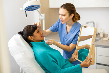Obraz premium Portrait of woman sitting on couch and looking at mirror during appointment with doctor at cosmetology clinic