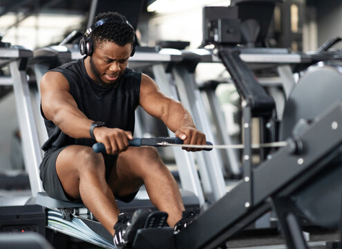 Motivated young black sportsman with wireless headset having workout on rowing machine. Handsome african american man bodybuilder training body on modern block exerciser in gym, copy space
