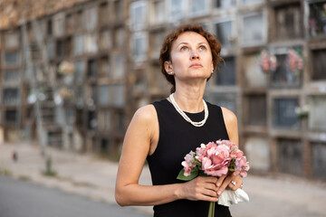 Fototapeta premium Sad woman in black mourning dress and pearl necklace standing in front of columbarium in old graveyard holding fresh flower bouquet, immersed in grief..