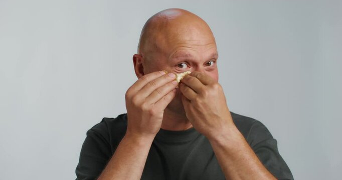 A bald man puts patches under his eyes. He smiles at the camera while standing in the studio background