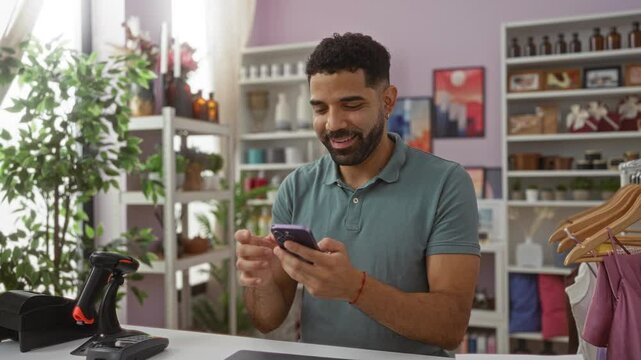 Young man celebrates with a fist pump while using a phone in a home decor store, surrounded by plants and shelves displaying various decorative items