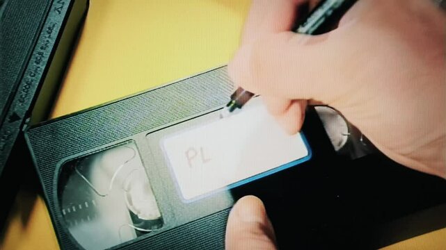 Real capture: hand writes Play Me with black marker on weathered VHS tape label. Analog nostalgia. Personal touch meets retro technology in evocative vintage moment.
