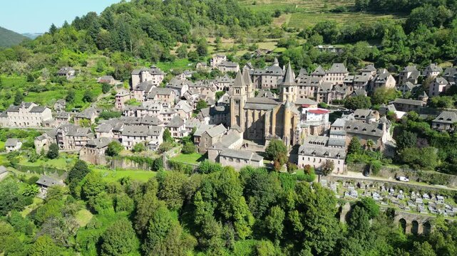 Drone aerial view in France countryside small old medieval town and a cathedral surrounded by a green mountain forest circling around in Conques