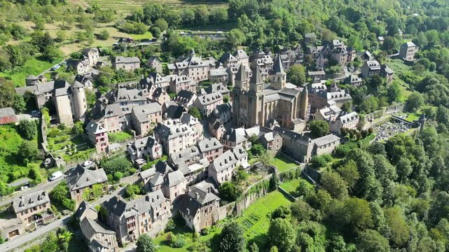 Drone aerial view in France countryside small old medieval town and a cathedral surrounded by a green mountain forest flying away in Conques