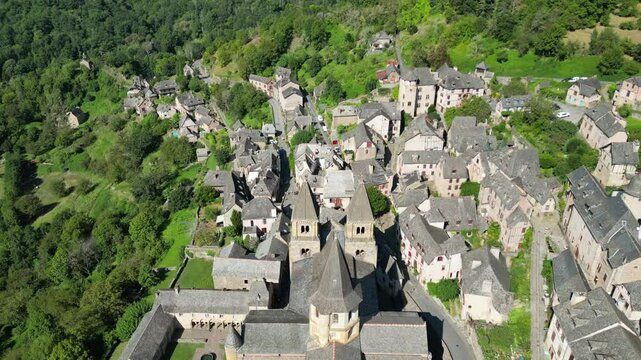 Drone aerial view in France countryside small old medieval town and a cathedral surrounded by a green mountain forest flying over in Conques