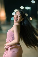 Young woman with long hair joyfully posing at night, wearing elegant dress, with soft lights in the background creating a vibrant urban atmosphere.