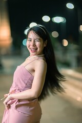 Fototapeta premium Joyful young woman in elegant dress smiles and poses playfully outdoors at night, illuminated by city lights in a vibrant urban setting