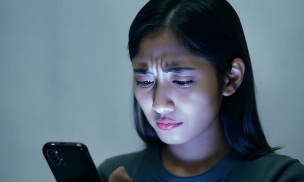 An Asian young woman holding her phone while gazing at the screen with a look of stress and concern. Her furrowed brow and tense expression conveys the negative impact of social media on mental health