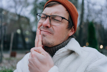Young man wearing glasses and winter clothes touching his jaw and suffering from a strong toothache...
