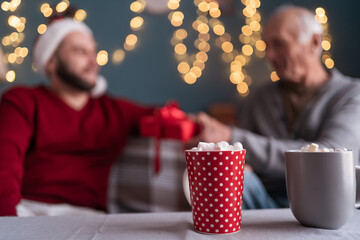 Son giving gift to retired father visiting for christmas sitting on sofa with cups of cocoa with marshmallows in foreground, selective focus