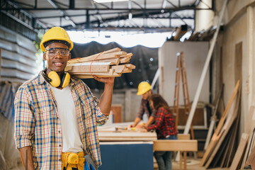 Smiling construction worker in a yellow hardhat and plaid shirt holding wood planks on his shoulder. Ideal for carpentry, woodworking, and craftsmanship in furniture workshop, National Carpenters Day