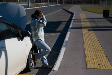 Sad Caucasian woman calling on phone while standing near car with open hood. 