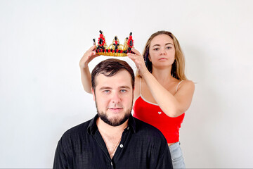 A woman places a golden crown with red and black jewels on a man's head, symbolizing patriarchy and...
