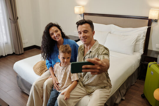 Family of three taking selfie in cozy hotel room with a bed and luggage in the background sharing happy moment together