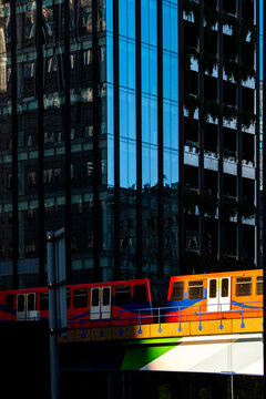 A vibrant train moves along tracks beneath towering skyscrapers, casting reflections in the early morning light, highlighting the modern city architecture.