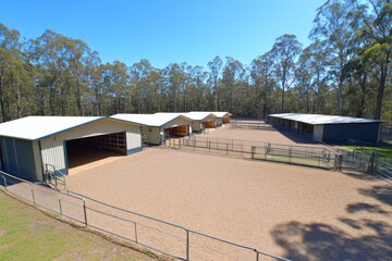 Fototapeta premium A row of horse stables with an open riding arena, surrounded by trees.