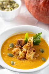 In a white ceramic bowl, a creamy pumpkin soup garnished with croutons, parsley and pumpkin seeds. To the side, on a textured white background, a pumpkin.
