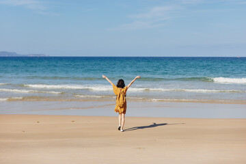 Joyful woman in a sunny yellow dress standing on a sandy beach with arms raised in celebration