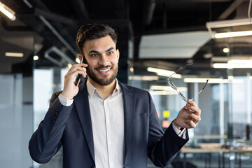 Confident businessman in office talking on phone, smiling while holding glasses. Represents modern...