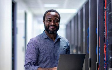 Portrait of a focused IT technician in a modern office, holding a laptop and a network cable, smiling at the camera.