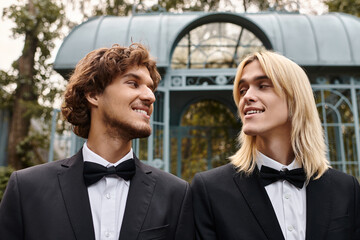 Two young men dressed in stylish black suits share a joyful connection in a beautifully landscaped garden.