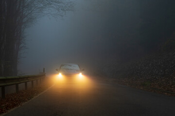 
A car with glowing headlights drives along a foggy forest road at dusk, creating a moody and...