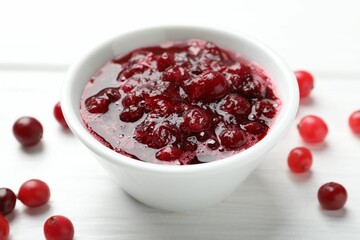 Tasty cranberry sauce in bowl and berries on white wooden table, closeup