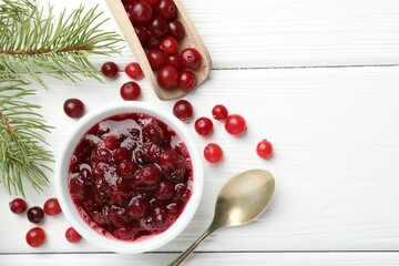 Tasty cranberry sauce in bowl, berries, spoon and fir branches on white wooden table, top view. Space for text