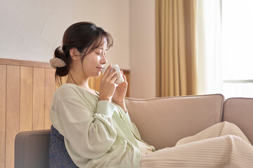 ソファーに座り温かい飲み物を飲みながらリラックスしている若い女性。