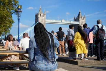 A woman in denim sits on a bench near Tower Bridge in London, surrounded by a diverse crowd enjoying a sunny day. Perfect for concepts of travel, tourism, urban life, and multiculturalism