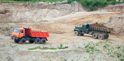 Two trucks are actively engaged in transporting materials at a construction site. The bright orange and green trucks move across the sandy quarry