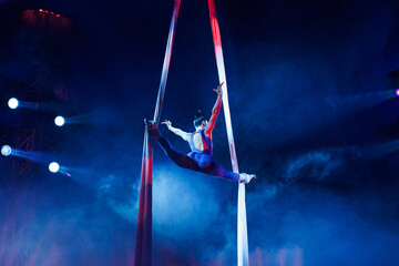 Aerialist performs a dangerous trick on a fabric high under dome of the circus under the spotlight.