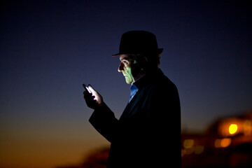 A man stands silhouetted against the dusk sky, intently focused on his smartphone. He wears a hat and is located near the shoreline, with distant lights twinkling in the background.