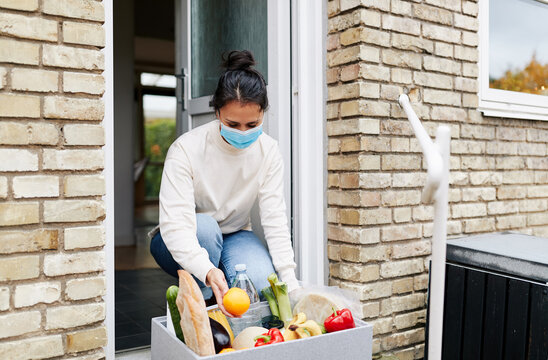 Woman in a blue mask receiving a grocery delivery. The box is filled with fresh organic vegetables and bread.