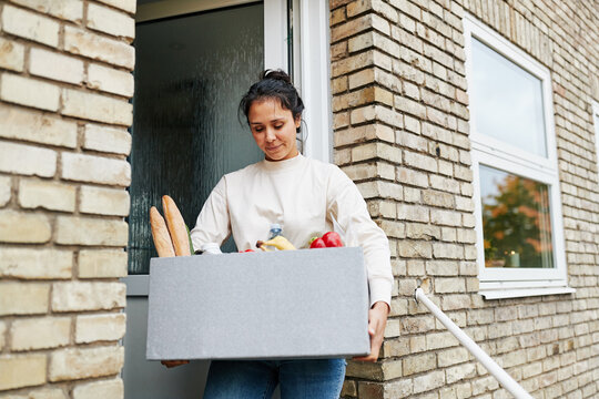 Smiling woman standing outside her front door, holding a grocery delivery box filled with fresh organic vegetables, bread and groceries.