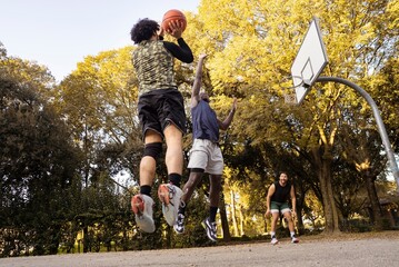 A basketball player jumps to shoot the ball as a defender leaps to block it, captured under bright sunlight on an outdoor park court with tall trees in the background