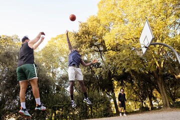 A basketball player jumps to shoot the ball as a defender leaps to block it, captured under bright sunlight on an outdoor park court with tall trees in the background