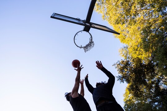 A low-angle shot of two basketball players reaching for the ball as it nears the hoop under clear blue skies on a bright and sunny day in a park setting