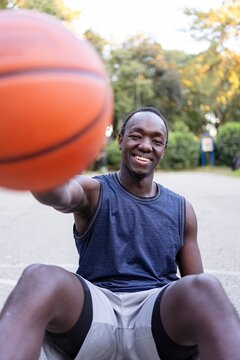 A basketball player sits on the court smiling and holding out the ball, surrounded by greenery and bathed in warm sunlight in a serene park