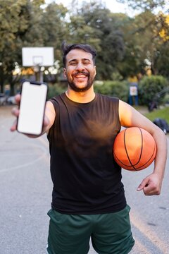 A basketball player seated on the court holding a ball and smiling warmly under bright sunlight surrounded by lush park greenery