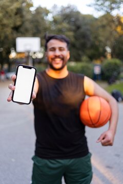 A basketball player seated on the court holding a ball and smiling warmly under bright sunlight surrounded by lush park greenery